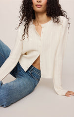 Z SUPPLY Medina Pointelle Cardigan in Cloud ivory, featuring button front, cable-knit texture, and long sleeves, styled with blue jeans on a seated model.