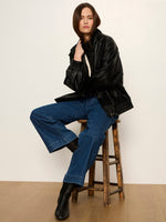 Sanctuary Alana Jacket in black faux leather, oversized fit with elastic hem, worn by model seated on wooden stool with wide-leg jeans