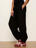 Sanctuary Micro Balloon Pant in black, side view on model with elastic waistband, draped harem-style silhouette, paired with burgundy sneakers.
