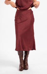 Sadie Ceremony Midi Skirt in dark wine satin, front view on model paired with knee-high brown leather boots