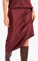 Sadie Ceremony Midi Skirt in dark wine satin, asymmetric wrap silhouette with draped hem, styled with brown knee-high boots