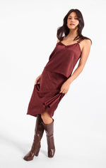 Sadie Ceremony Midi Skirt in dark wine satin, worn by model with matching cami top and brown knee-high boots, front view