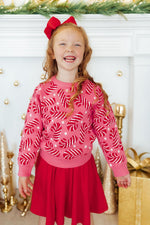 Mila Candy Canes Sweater in pink with red and white candy cane pattern, worn by young girl with red skirt in holiday setting.