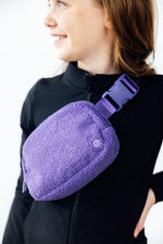 Mila Purple Fleece Belt Bag worn crossbody by a model in a black zip-up, featuring sherpa texture and purple buckle strap.