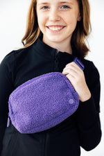 Mila Purple Fleece Belt Bag held by a smiling young model wearing a black zip-up, showing fluffy boucle texture and zipper detail.