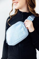 Mila Light Blue Fleece Belt Bag worn crossbody by smiling model in black zip-up, featuring sherpa texture and blue buckle strap.