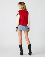 Steve Madden Rooney Jacket back view on model — red body, cream leather sleeves, black stripe cuffs, worn with denim mini skirt and boots