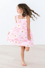 Girl wearing Mila & Rose Ruffle Cross Back Dress in pink with rainbow and sun pattern, shown mid-twirl