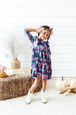 Mila Vintage Garden Ruffle Twirl Dress in navy floral print, worn by young girl with white cowboy boots and bow hair clip, fall setting