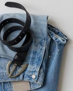 Free People WTF Noa Belt in black leather with silver western buckle and punched holes, styled on a folded light-wash denim jacket flat lay.
