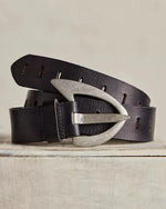 Free People Wtf Noa Belt in black, coiled on a wooden surface, featuring a pointed silver-tone buckle and slim leather strap with slots