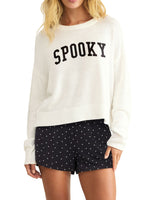 Z Supply Spooky Sweater in cream/sandstone knit with bold black collegiate lettering, worn by model with black polka dot shorts, front view.