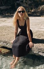 Z SUPPLY Johanna Midi Dress in black on a blonde model sitting on coastal rocks with feet in water, wearing cat-eye sunglasses.