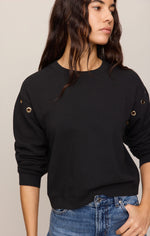 Z SUPPLY Axel Grommet Sweatshirt in black with gold grommet hardware along sleeves, worn by a model with blue jeans, front view.