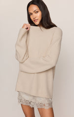 Z SUPPLY Chill Mode Sweater in Frosted Pearl, an oversized cream knit mock-neck pullover worn by a model with a lace mini skirt, front view.