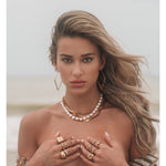Sahira Lori Hoops in gold — diamond-shaped geometric gold earrings worn by a model at the beach with layered necklaces and rings.