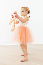 Young girl wearing Mila Ballet Tank Tutu Dress in soft peach-blush with floral bodice and tulle skirt, holding ballet shoes