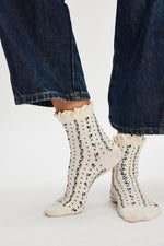 Free People Rosebud Waffle Knit Ankle socks in ivory with blue floral print and lace ruffle cuff, worn with dark wide-leg jeans.