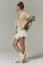 Free People Chelsea Jacket in Oatmeal worn by model with white pleated mini skirt and retro sneakers, side profile view.