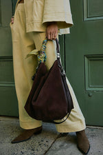 Free People Suzie Suede Slouch Tote in Espresso, a dark brown slouchy hobo bag with leather handle tied with a silk scarf, held by a model in a cream suit.